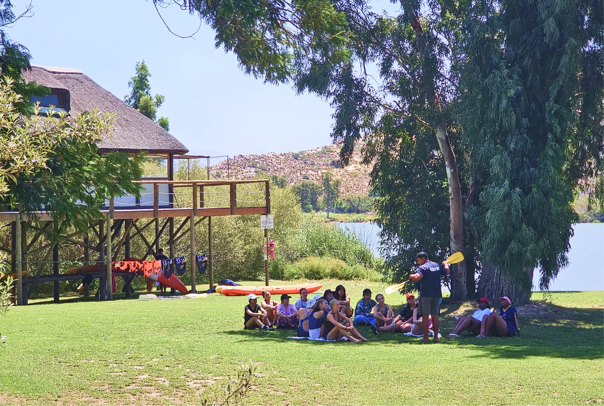 Large group seated on a riverside lawn during an outdoor briefing