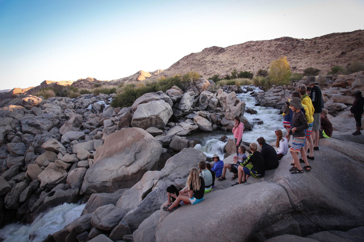 Group resting on rocks beside a river rapid at golden hour